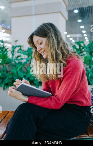 Young woman writing notes and journaling in a gray notebook with a pen, sitting on a bench, expressing positive emotions and a thoughtful, focused mindset Stock Photo