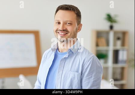 Portrait of smiling bearded man in casual wear standing at workplace in modern office or classroom. Stock Photo