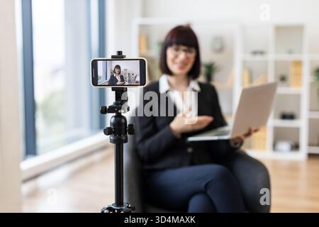Caucasian businesswoman in her 30s filming vlog content with smartphone and laptop on desk, engaging with her audience in modern professional workplac Stock Photo