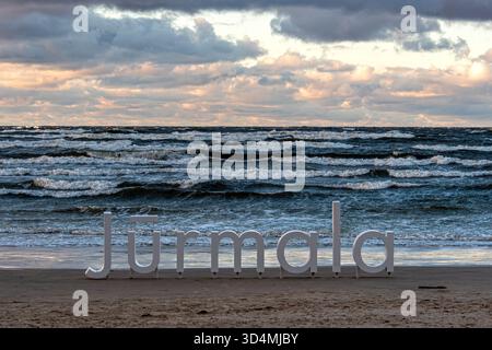 Jurmala sign stands on the beach as waves crash and the sun sets over the sea. Stock Photo
