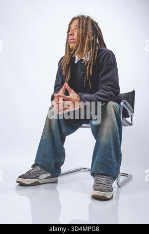 Young man with dreadlocks sitting on a chair, hands clasped, looking thoughtful Stock Photo