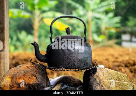 Horizontal close-up shot of an old, blackened cast iron or aluminum kettle (teapot) being heated on a traditional open fire setup, supported by rocks Stock Photo