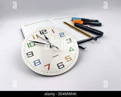 A still life composition centered around a clock face, a notebook, and various writing/marking tools. Stock Photo