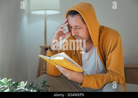 Relaxed adult male wearing yellow hooded shirt focused on reading a yellow book in a warm, comfortable living room setting, casual lifestyle moment. S Stock Photo