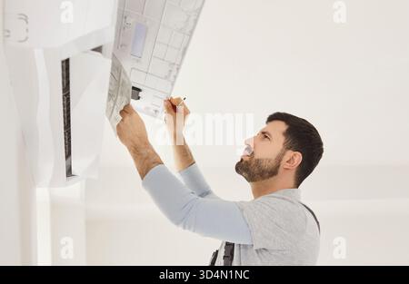 Man working, air conditioner unit mounted on wall, holding instructions, looking intently, focused  Stock Photo