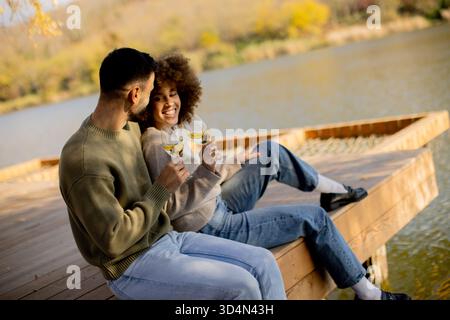 Two people sit together on a wooden dock, smiling and holding glasses of sparkling drink. The sun sets behind them, creating a warm glow. Stock Photo