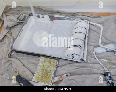 Worker pouring white paint into a paint tray covered with a plastic sheet, preparing to paint walls with a paint roller Stock Photo
