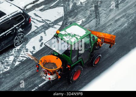 Above view small green tractor orange snow plow salt spreader clears icy street during winter in city. Fresh tire marks footprints on gray asphalt Stock Photo