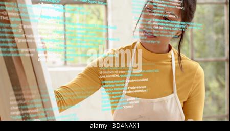 Reaching woman wearing mustard turtleneck and apron tracing code overlay on pane at home studio Stock Photo