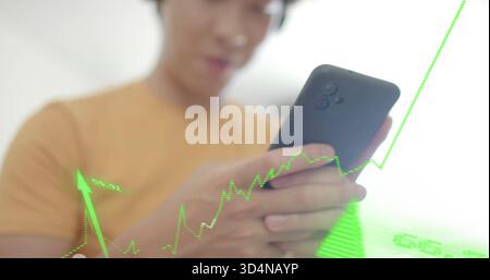 Holding dark-cased phone, Asian man in mustard shirt in bright room viewing green financial overlay Stock Photo