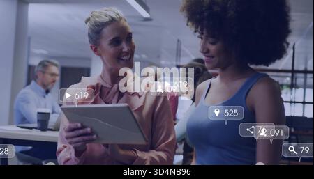Smiling two women holding cracked tablet viewing social overlays in office, pink blouse blue top Stock Photo