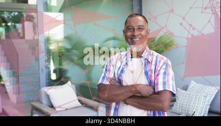 Standing senior man crossing arms and smiling in sunroom, with plaid shirt and cushioned seating Stock Photo