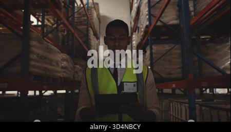 Inspecting man holding tablet checking stock in warehouse aisle, in neon vest, bulk containers Stock Photo