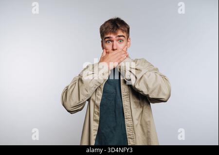 A young man stands with wide eyes and an open mouth, covering it with his hands, showing shock or disbelief in a neutral setting. Stock Photo