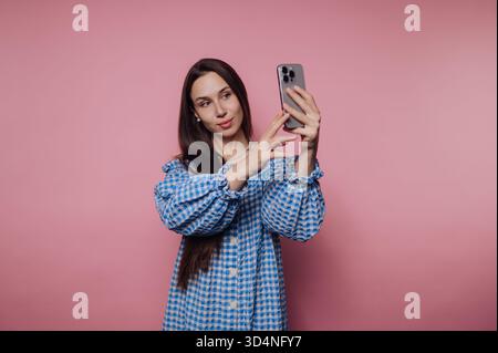 A woman smiles while capturing a selfie with her smartphone in front of a bright pink wall. Her long hair and checkered dress add to the joyful atmosp Stock Photo