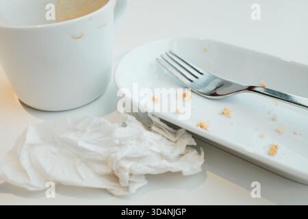 Empty cup with coffee foam, white plate with crumbs, fork, and crumpled napkin after a meal. Stock Photo