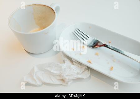 Empty cup with coffee foam, white plate with crumbs, fork, and crumpled napkin after a meal. Stock Photo