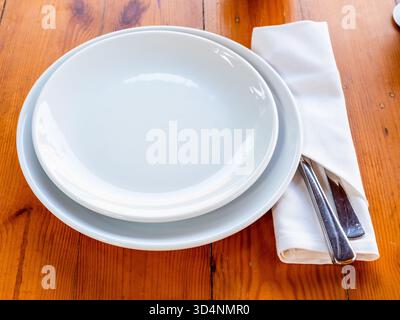 empty white plate in bowl and knife and fork in napkin served on wooden table Stock Photo