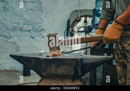 Hands of craftsman in gloves with iron hammer align iron part on stone. Vintage construction tool. Preparation for welding work. Stock Photo