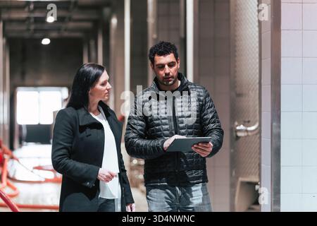 Two professionals engage in a conversation while reviewing a tablet in a contemporary industrial setting. Stock Photo