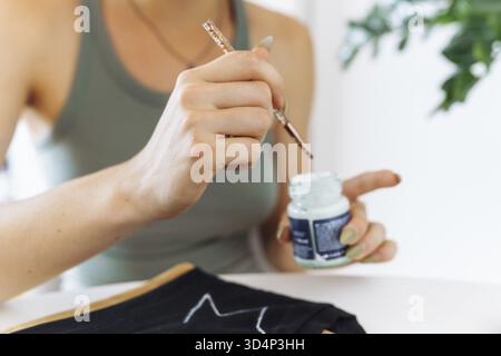Young woman draws picture on clothes with paints, handmade craft work. An artist uses brush to trace lines design on clothing. close-up Stock Photo
