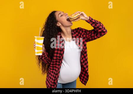 Funny Young Pregnant Lady Excitedly Enjoys a Big Burger and Drink Against a Yellow Backdrop Stock Photo
