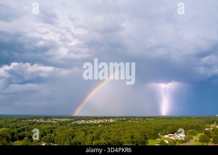 Vibrant rainbow spans sky as storm brews, lightning striking in distance over lush greenery. Stock Photo