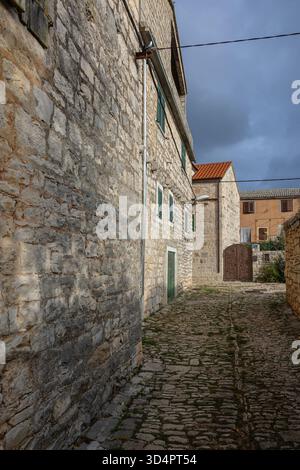 A sunlit, narrow street winds between historic stone houses with wooden shutters and tiled roofs, capturing a tranquil, rustic village atmosphere and Stock Photo