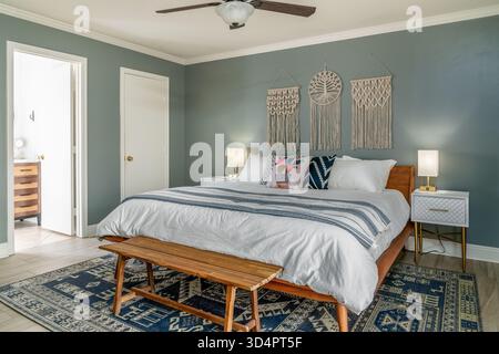 Modern boho bedroom interior. Bed, pillows, blue wall, macrame hangings, lamps, nightstands. Wood bench and rug. Stock Photo