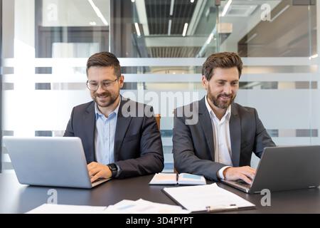 Two smiling bearded businessmen in suits working on individual laptops at a bright modern office desk, focused on planning, collaboration and productive teamwork tasks Stock Photo