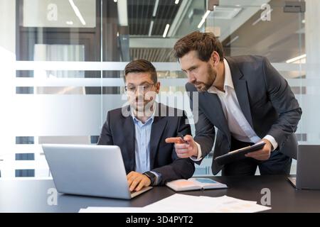 Two professional businessmen collaborating while looking at a laptop and pointing to the screen, reviewing reports and discussing new strategies during a corporate meeting in a modern office Stock Photo