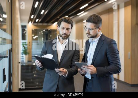 Two businessmen in a modern office corridor review documents on a clipboard and tablet, collaborating on strategy and communication to plan a project and ensure successful partnership Stock Photo