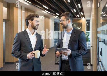 Two professional businessmen discussing a new project, standing in a contemporary office corridor, one holding a coffee cup and the other a digital tablet while exchanging ideas Stock Photo