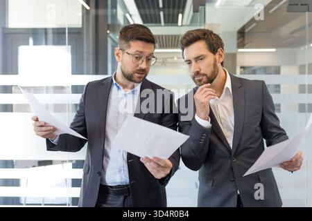 Two businessmen are collaborating while reviewing documents, analyzing important financial records and business calculations in an modern office environment, planning next steps Stock Photo