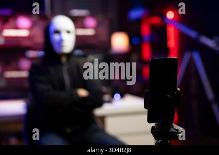 Close up of masked man using phone to film video, doing espionage campaign from hidden bunker. Focus on smartphone used by scammer wearing anonymous mask to record demands from secret location Stock Photo