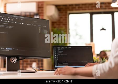 Closeup of female freelancer uses multiple device screens, writing programming scripts at home. Young adult developer building website and reviewing code in modern brick wall apartment. Stock Photo