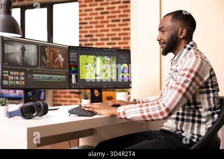 Male post production artist edits content using dual screen setup at home. African american video editor makes creative decisions while organizing footage and timelines on desktop pc. Stock Photo
