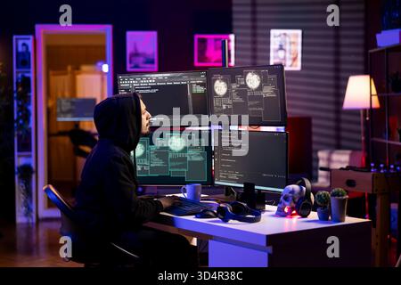 Hooded hacker in neon lit lair using PC to gain unauthorized network access, targeting unpatched connections. Rogue engineer at home bypassing security and exploit servers for stolen data profit Stock Photo
