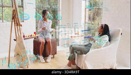 Painting two women collaborating near easel in bright art studio, in overalls, holographic overlays Stock Photo