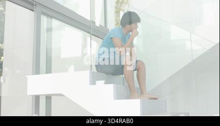 Sitting man in blue tee and denim-shorts on white floating stairs by glass with foliage reflections Stock Photo