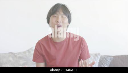 Sitting Asian man wearing red T-shirt speaking and gesturing on sofa in lounge, with pillows Stock Photo