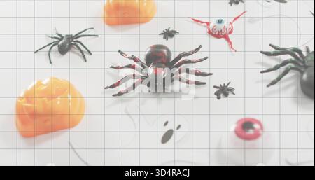 Sitting glossy black-and-red plastic spider on white grid mat at table, with pumpkins and eyeballs Stock Photo