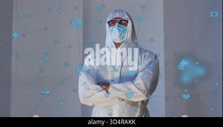 Standing man in hooded coverall at clinical panels, wearing mask, goggles, arms crossed, blue icons Stock Photo