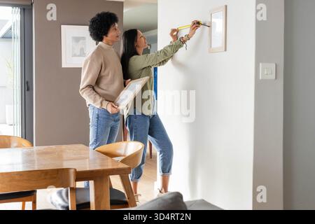 Diverse couple measuring and hanging picture frame on wall by dining table with yellow tape measure Stock Photo