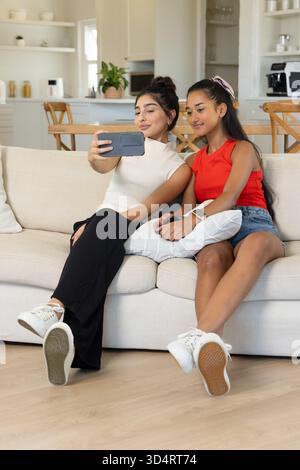 Taking selfie Diverse female friends leaning on sofa in open-plan living room, with smartphone Stock Photo
