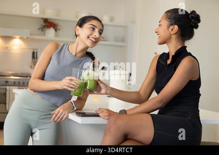 Clinking glasses Diverse female friends laughing at modern kitchen island, with green smoothies Stock Photo