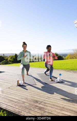 Practicing diverse female friends doing low lunge yoga on deck by lawn, with mats towels bottles Stock Photo