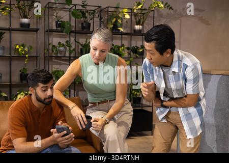 Diverse coworkers testing devices on brown couch in office lounge, with smartphone and speaker Stock Photo