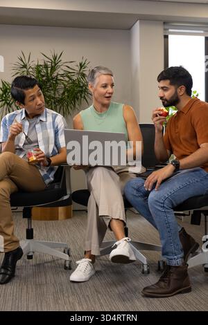 Snacking on fruit cup and apple, Diverse coworkers sitting in office lounge by plant with laptop Stock Photo