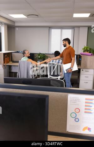 Shaking hands Diverse coworkers standing in open-plan cubicles, with computer monitors and yoga mat Stock Photo